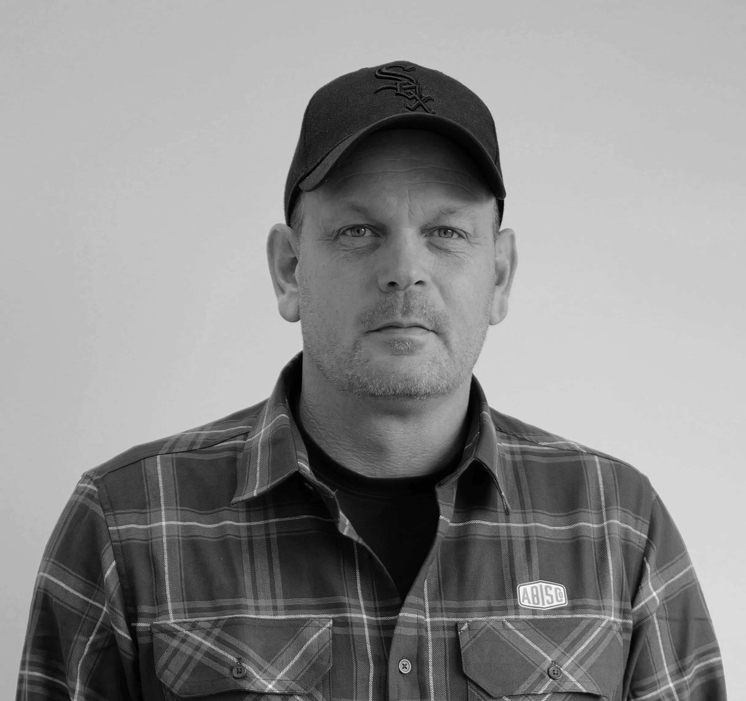Black and white portrait of a man wearing a cap with a sports logo and a plaid shirt, looking directly at the camera