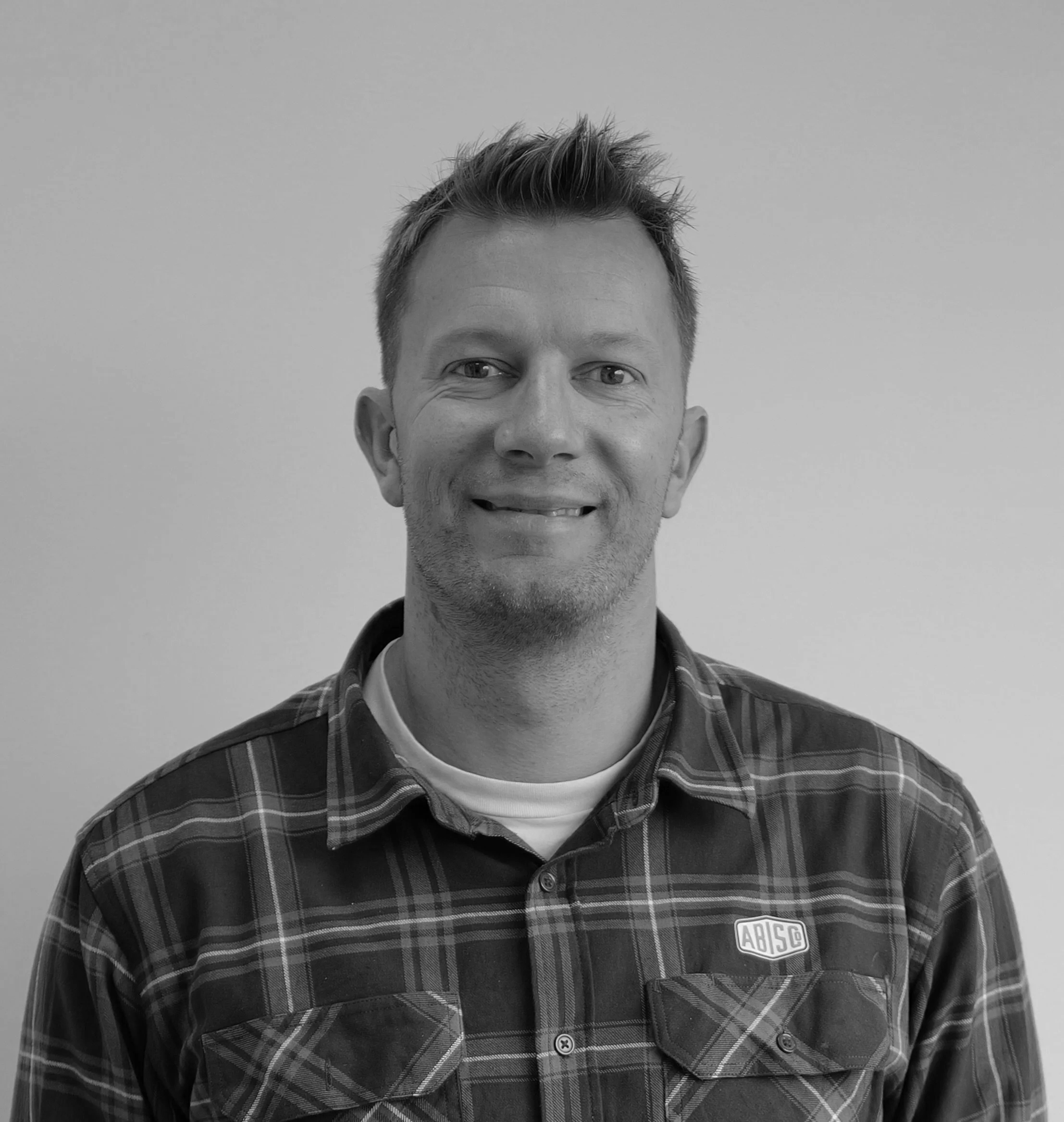 Black and white portrait of a smiling man with short hair, wearing a plaid shirt with an 'ABISCO' logo.