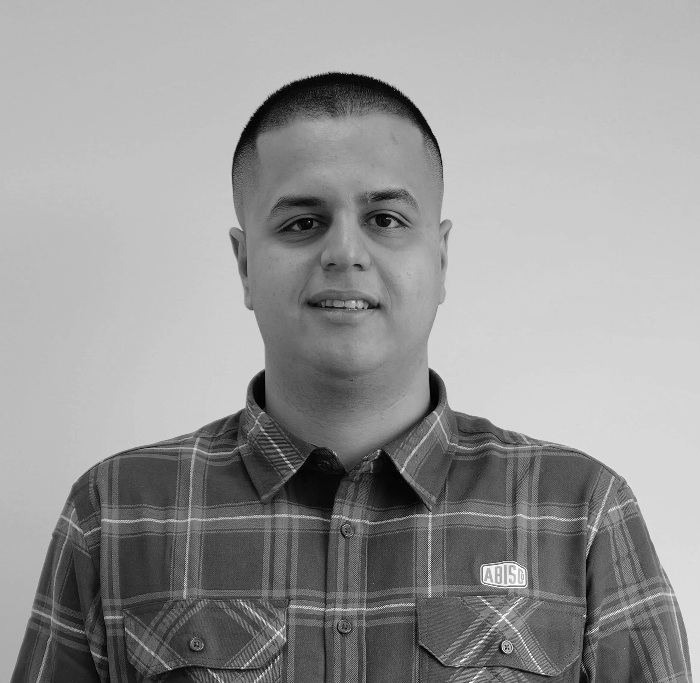 Black and white portrait of a young man with short hair, wearing a plaid shirt with an 'ABISCO' patch, standing against a plain background.