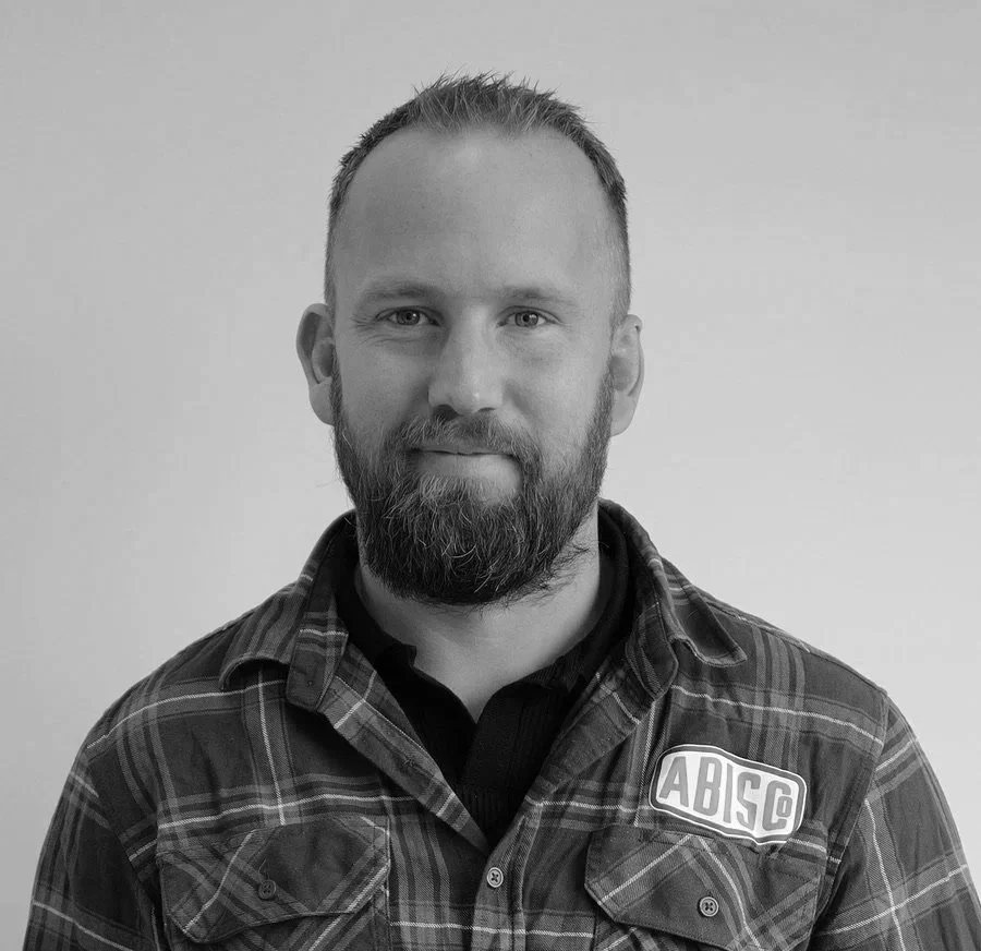 Black and white portrait of a man with a beard and short hair, wearing a plaid shirt with a patch reading 'ABSC'.