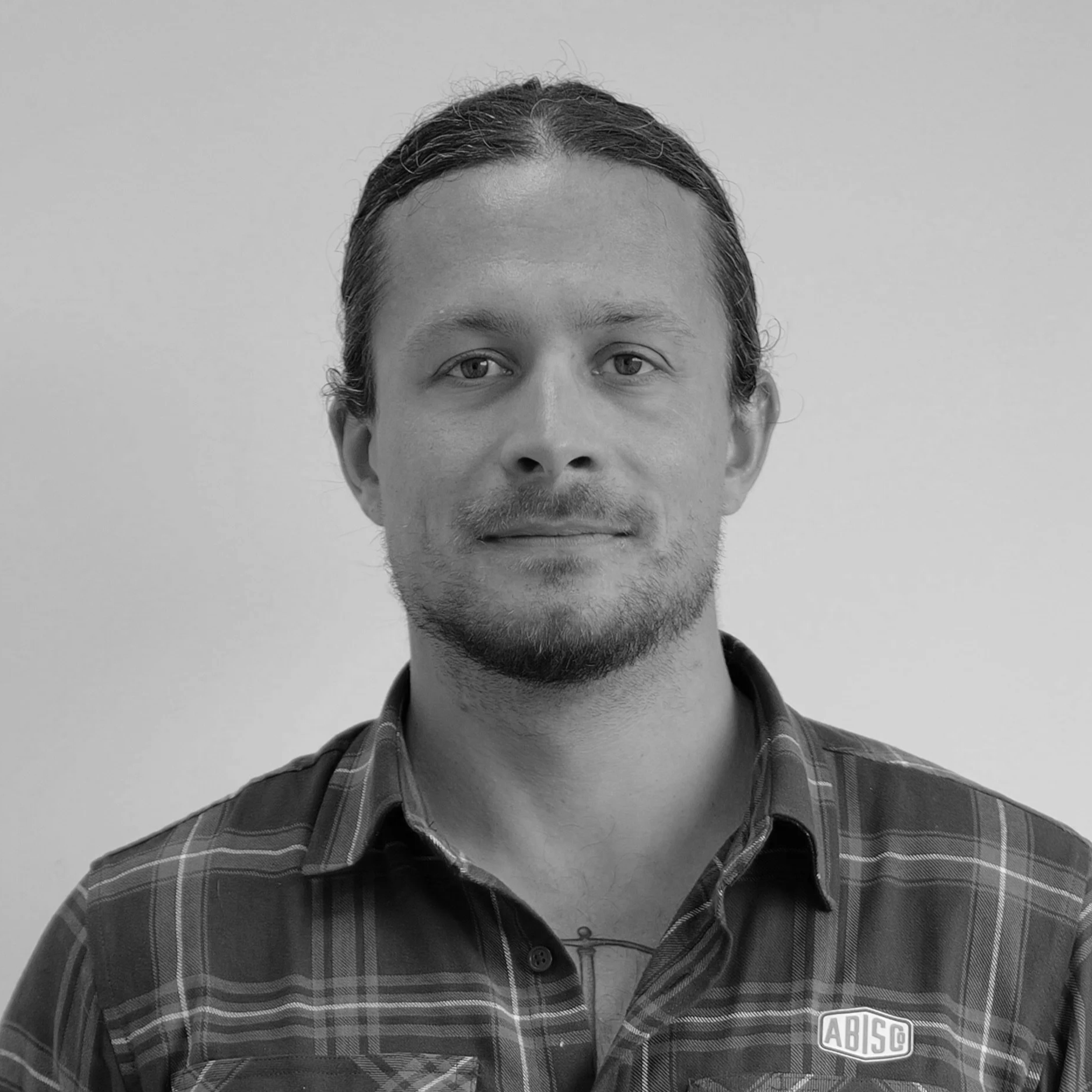 Black and white portrait of a young man with shoulder-length hair, slight beard, wearing a plaid shirt with a logo that reads 'ABISC'