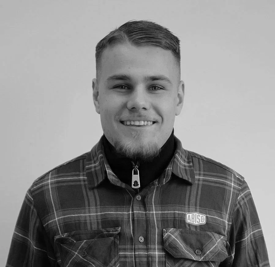 A young man with short hair, a beard, and light skin, smiling at the camera. He is wearing a plaid shirt with a logo on the left side and a zip-up jacket underneath against a plain background.