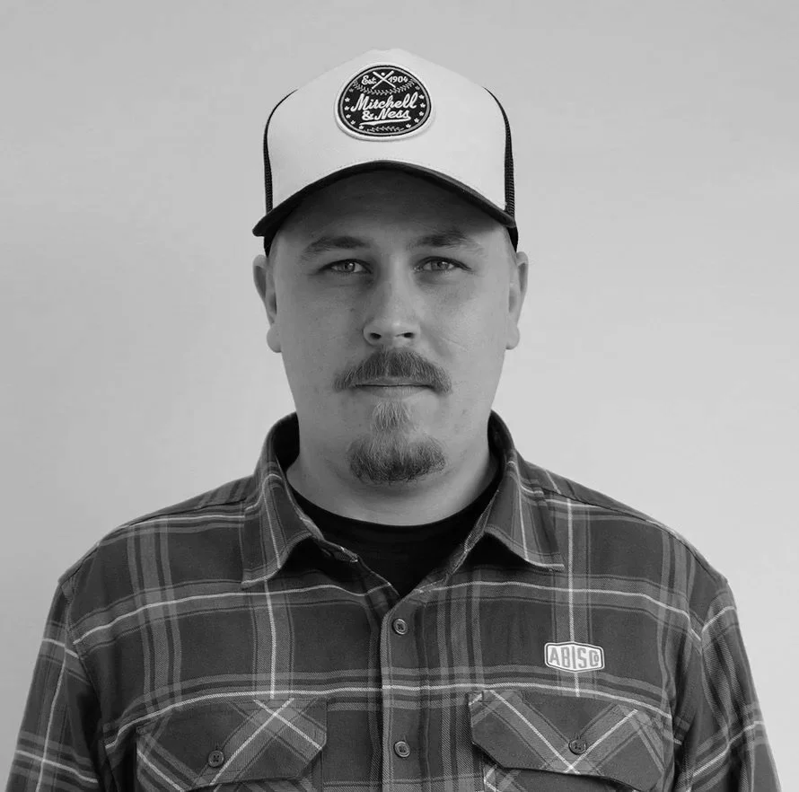 Black and white photograph of a man with a mustache and goatee, wearing a plaid shirt and a trucker hat with a logo.