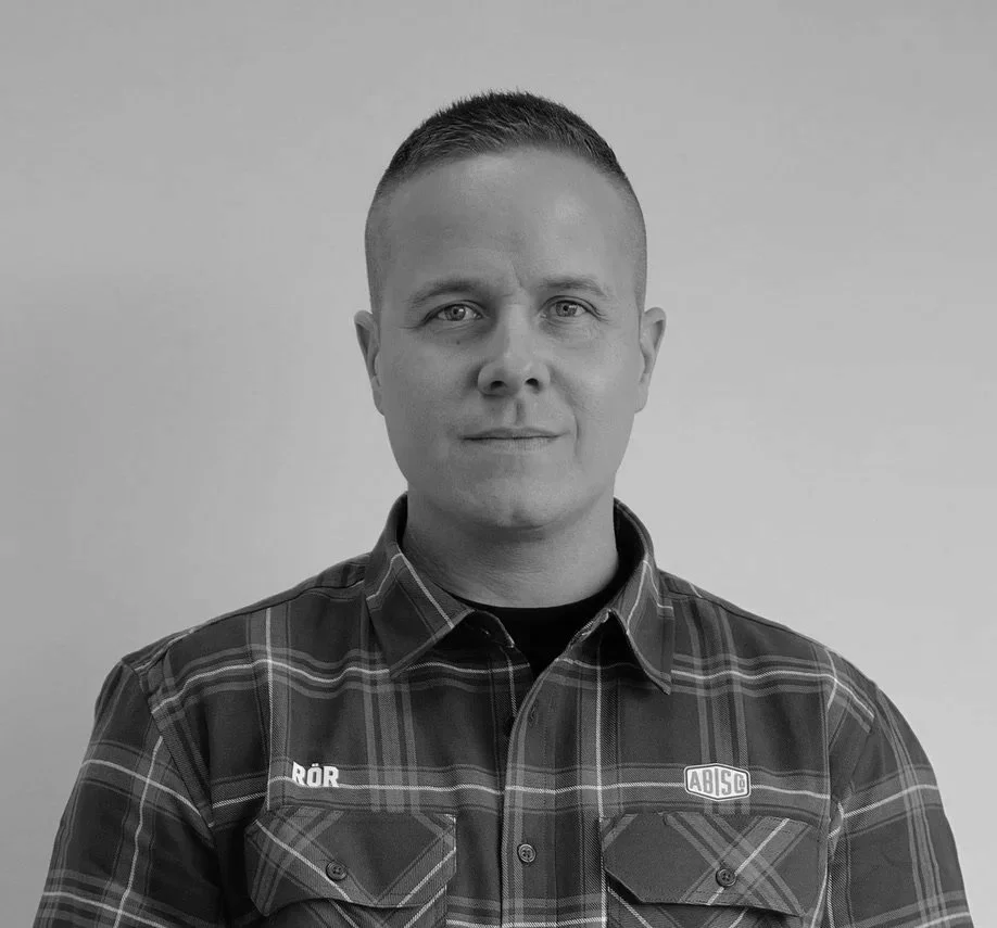A black-and-white portrait of a young man with short hair, wearing a plaid shirt with a logo on the chest, standing against a plain background.