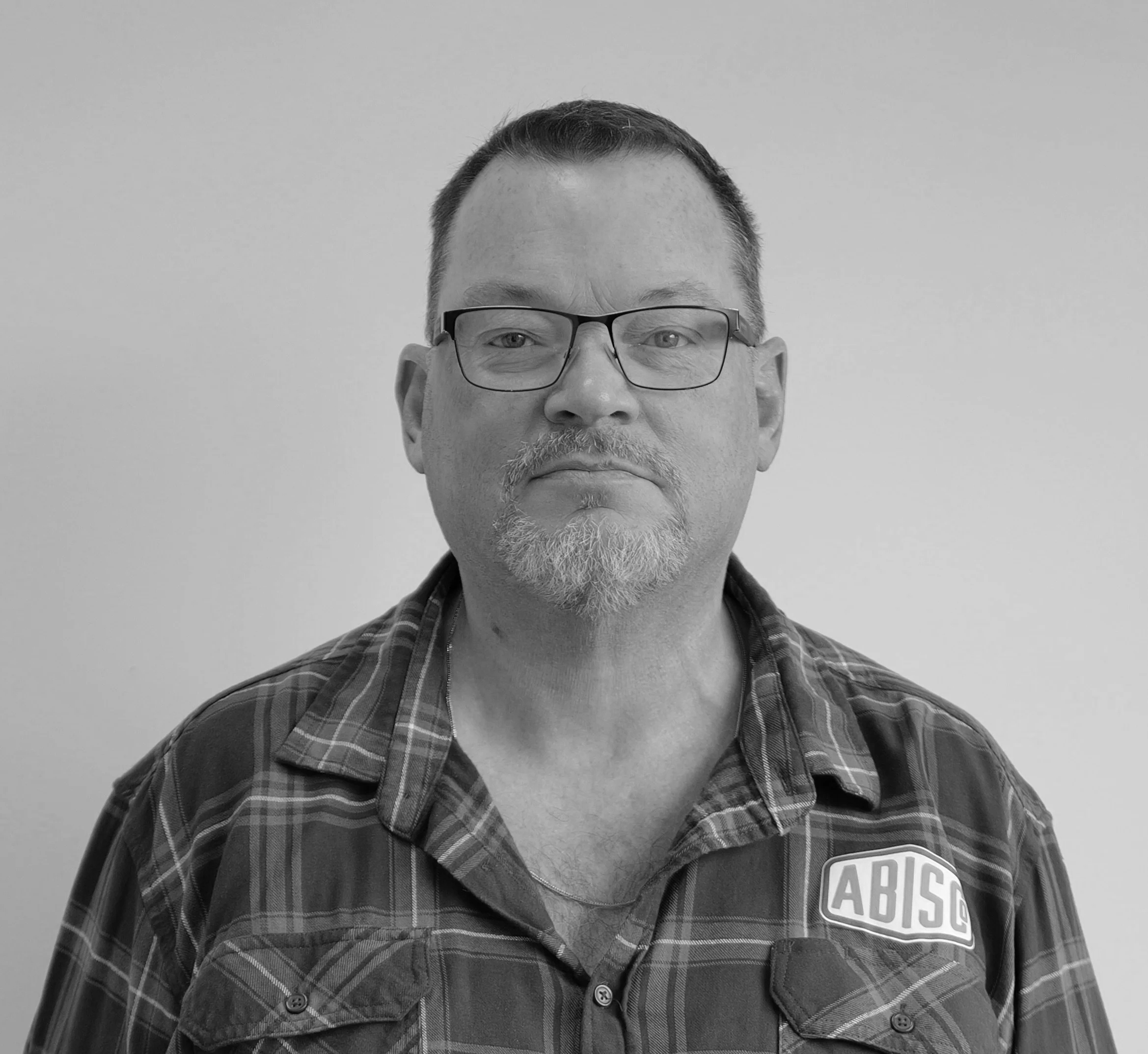 A middle-aged man with short dark hair, glasses, and a goatee, wearing a plaid shirt with an 'ABIS' patch, standing against a plain background.