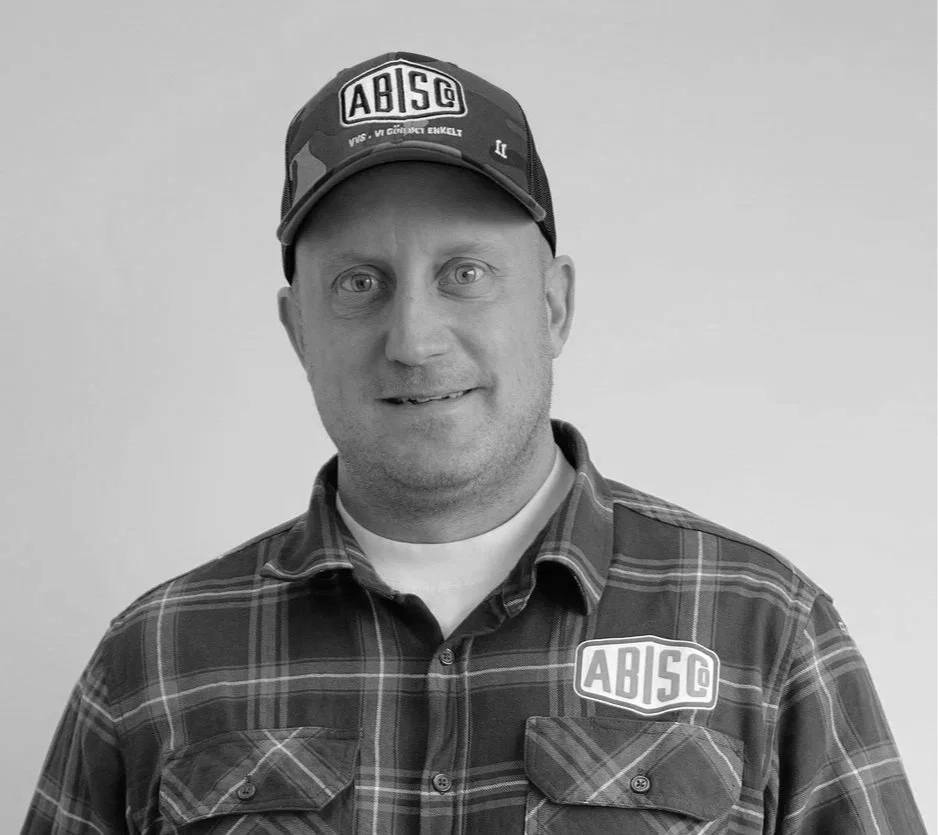 A man wearing a plaid shirt and cap with ABISG patch, smiling at camera against a plain background.