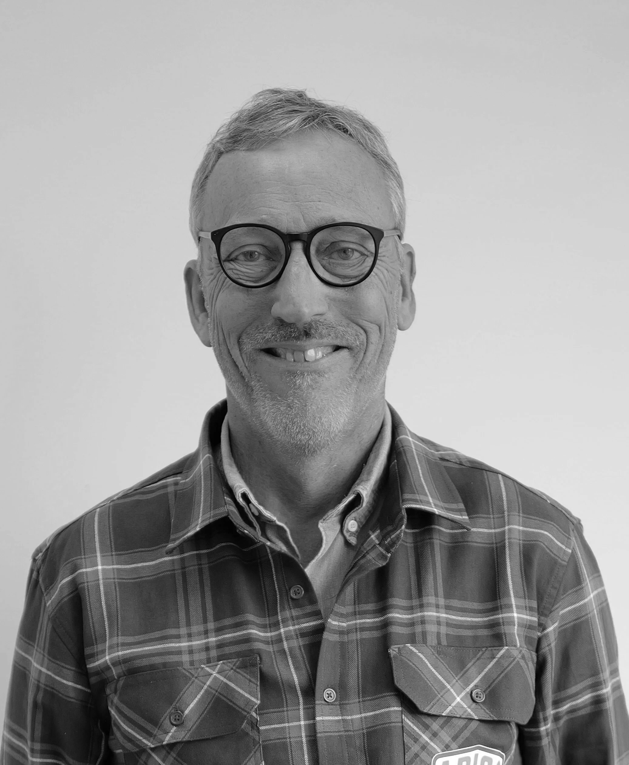 Black and white photo of a smiling middle-aged man with glasses, short hair, and dressed in a plaid button-up shirt with a logo on the chest, standing against a plain background.