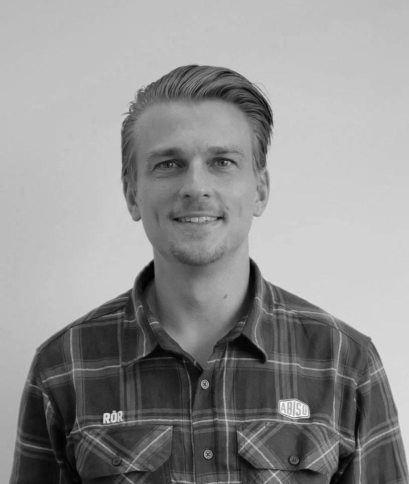 Black-and-white portrait of a smiling man with slicked-back hair, wearing a plaid shirt with patches on the chest, standing against a plain background.