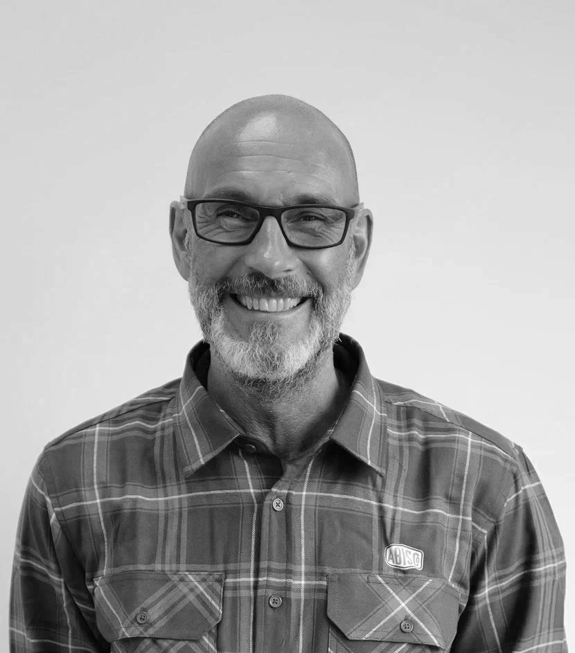 A smiling man with a bearded face, glasses, and a shaved head, wearing a plaid shirt, standing against a plain background.