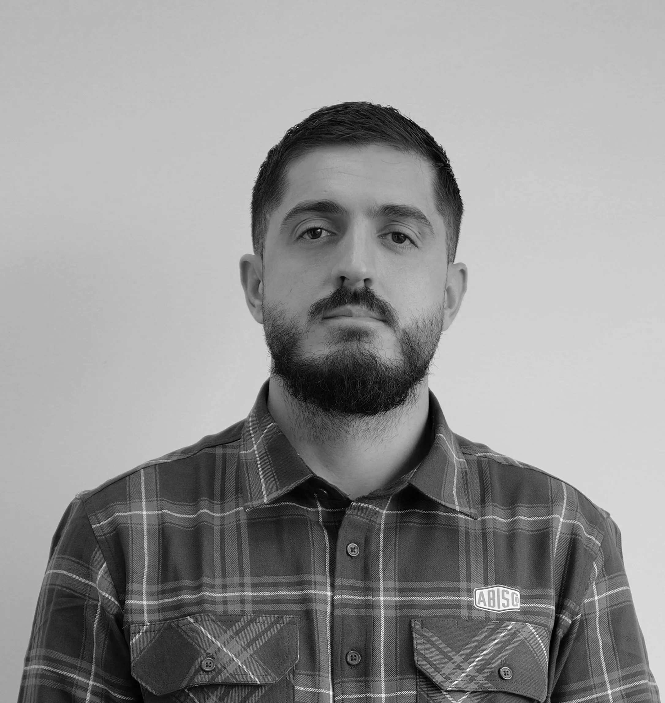 Black and white portrait of a man with short dark hair, a full beard, wearing a plaid shirt with a logo on the left chest, standing against a plain light-colored wall.