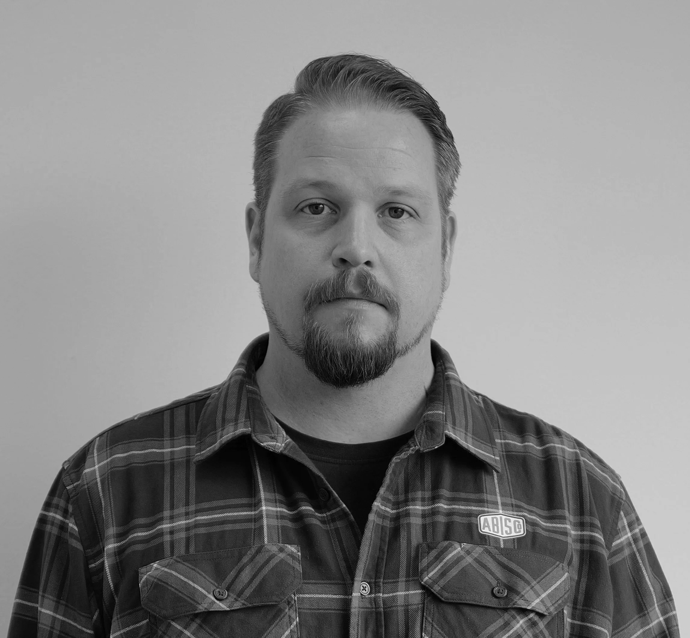 A man with light hair and a beard, wearing a plaid shirt with an 'ABISCO' patch, looking directly at the camera.