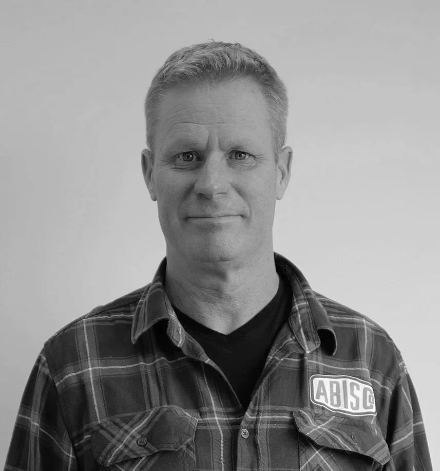 Black and white portrait of a middle-aged man with short hair, wearing a plaid shirt with an 'ABISCO' logo on the chest, standing against a plain background.
