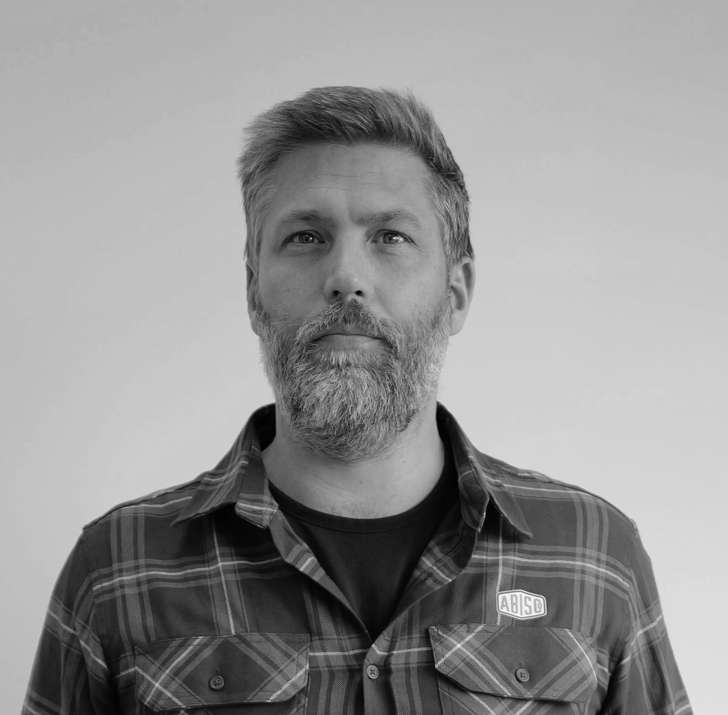 Black and white portrait of a middle-aged man with a beard and short hair, wearing a plaid shirt with an 'ABISCO' logo patch, facing the camera with a serious expression.