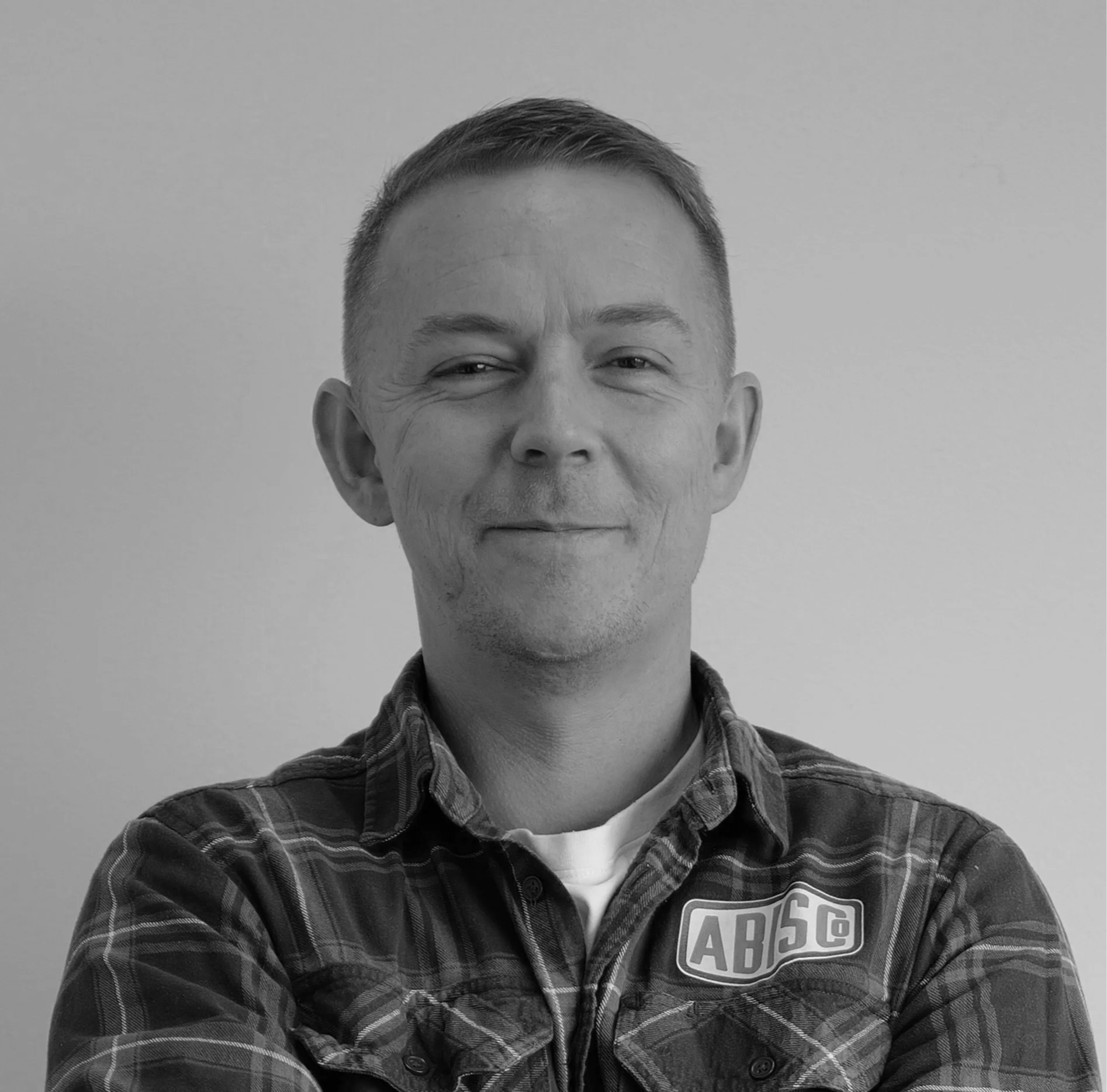 Black and white portrait of a middle-aged man smiling, wearing a plaid shirt with an 'ABS' logo, against a plain background.