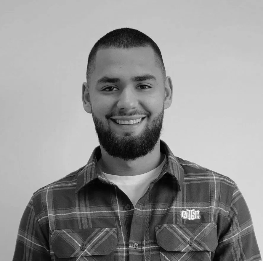 Black and white photo of a smiling man with a beard, short hair, wearing a plaid shirt with a logo on the chest, standing against a plain background.