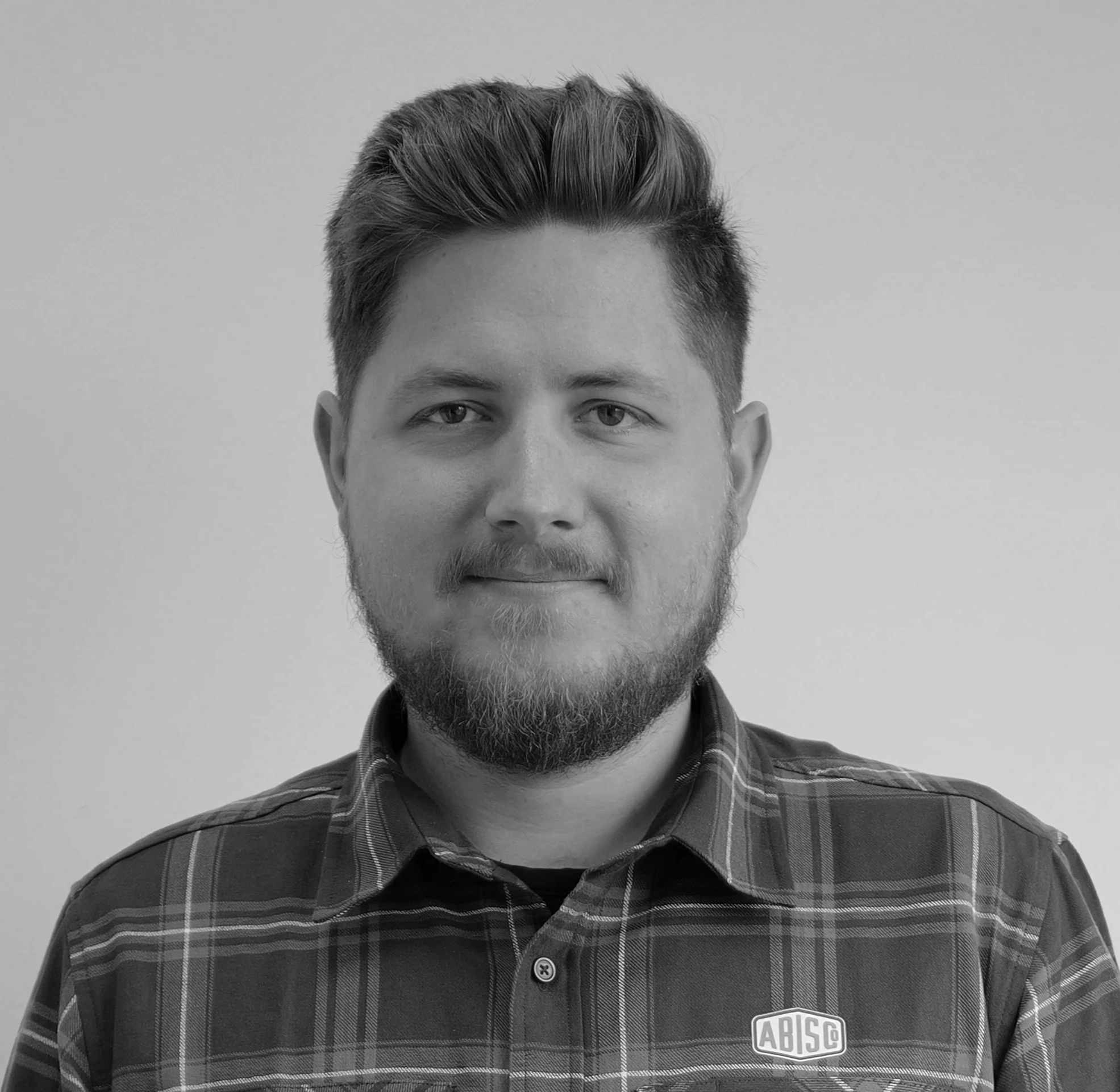 Black and white portrait of a young man with short, styled hair and a beard, wearing a plaid shirt with a company logo badge that reads 'ABISG'.