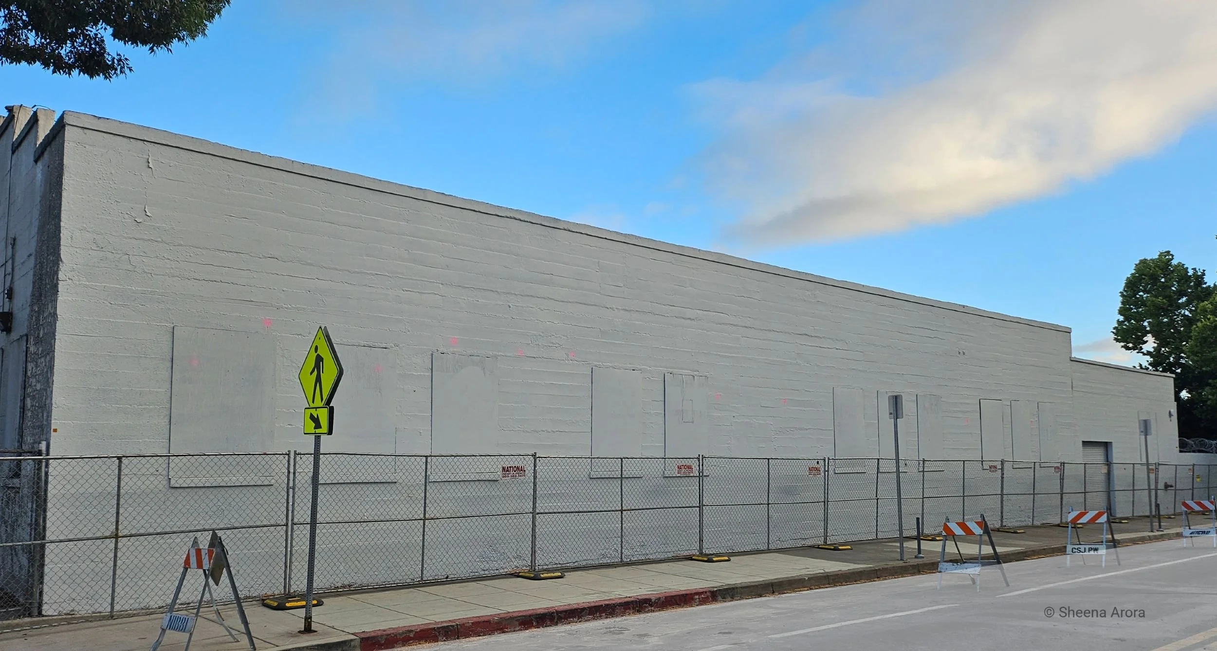 A long exterior concrete wall on 5th Street in San Jose, completely painted flat grey and surrounded by chain-link fencing where the IBM mural once stood, captured by Sheena Arora.