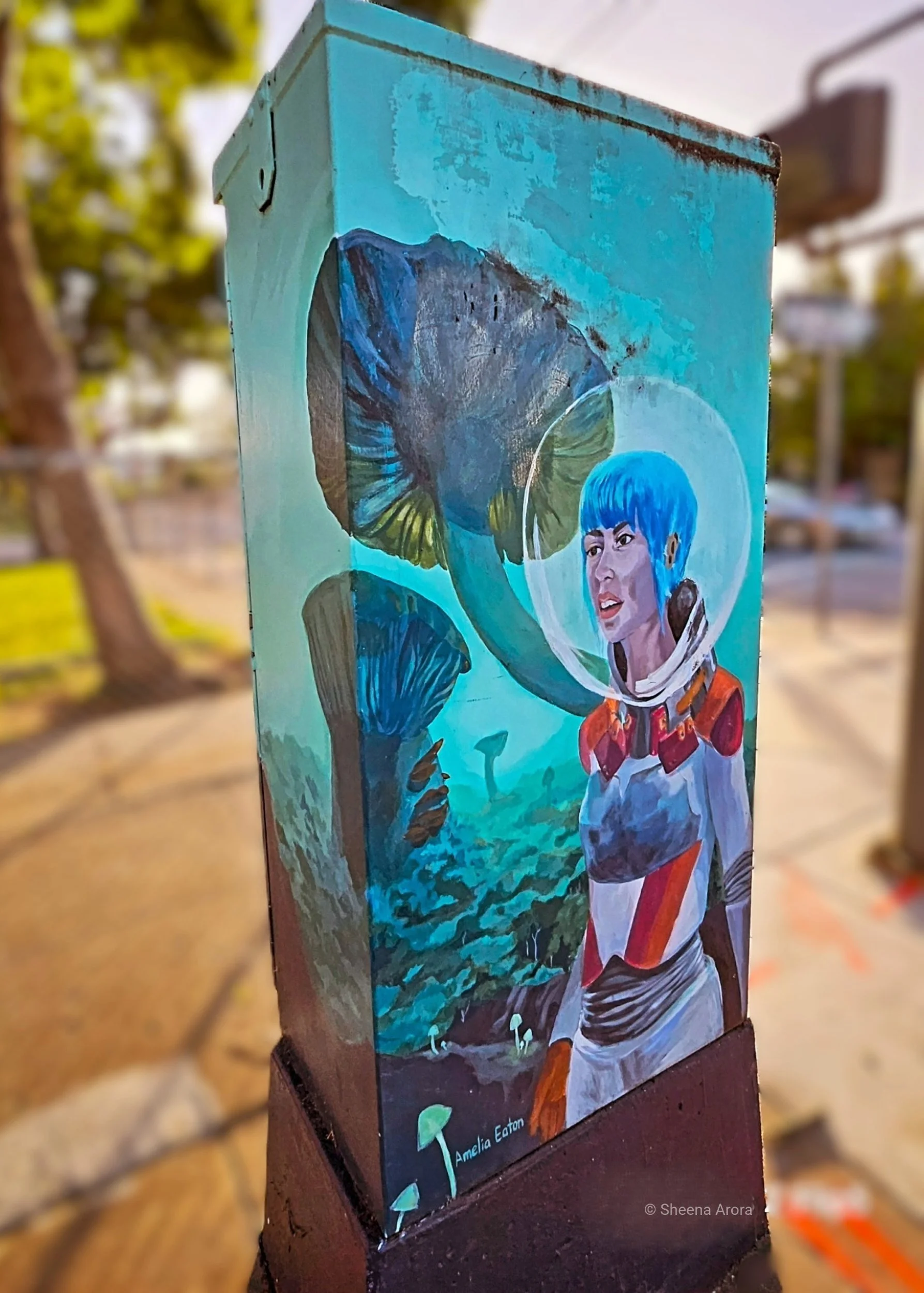 A street art mural painted on a tall metal utility box by Amelia Eaton depicts a space explorer with short bright blue hair wearing a clear bubble helmet and space suit. She stands among giant bioluminescent teal and blue mushrooms in an alien landsc