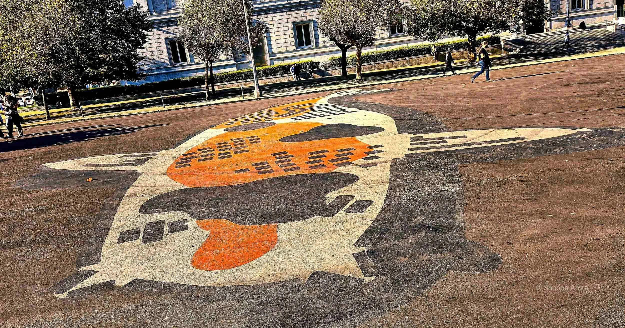 Large-scale street art mural of an orange, black, and white Koi fish painted by Jeremy Novy onto the asphalt pavement of Fulton Plaza in San Francisco, captured from a high-angle perspective with civic architecture in the background.