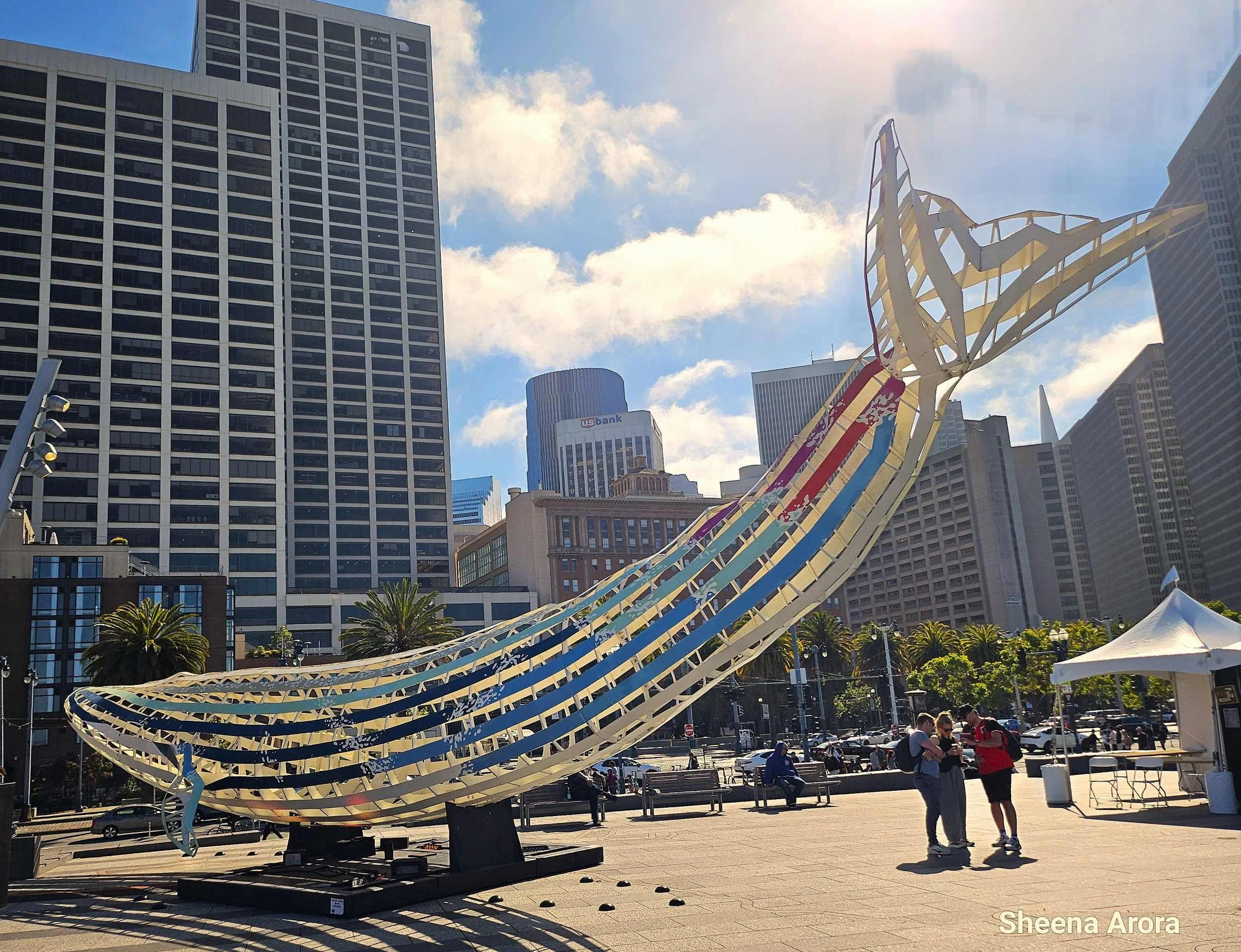 A large, skeletal industrial steel sculpture of a blue whale calf titled Echoes by Mathias Gmachl, featuring painted blue, wave-patterned, and red stripes along its body, set against the backdrop of palm trees and San Francisco skyscrapers at the Fer