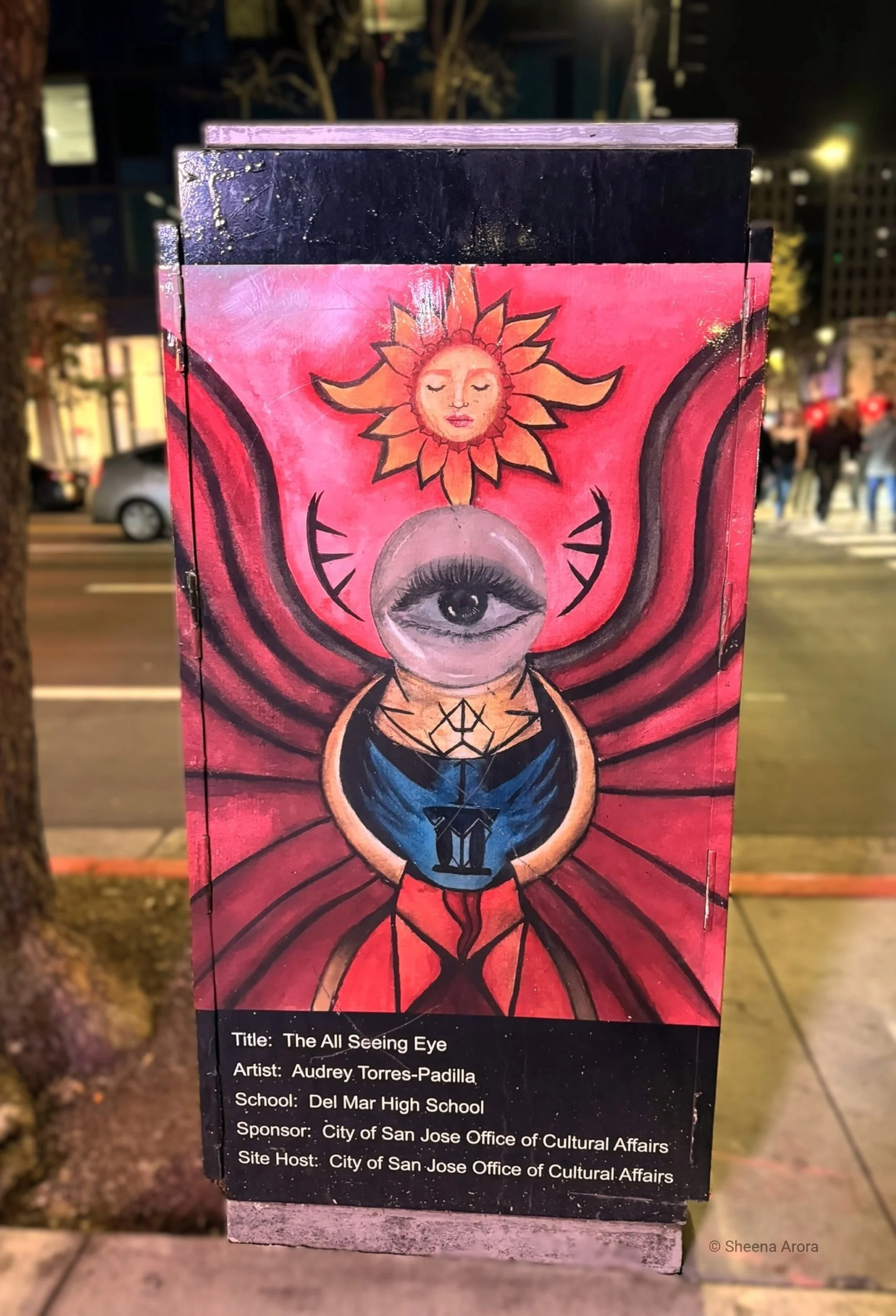 A vibrant, surreal street art painting on a utility box by Audrey Torres-Padilla showing a realistic human eye enclosed inside a crystal sphere. Radiant crimson bands and bright blue strokes curve around the center beneath a stylized, glowing sun fac