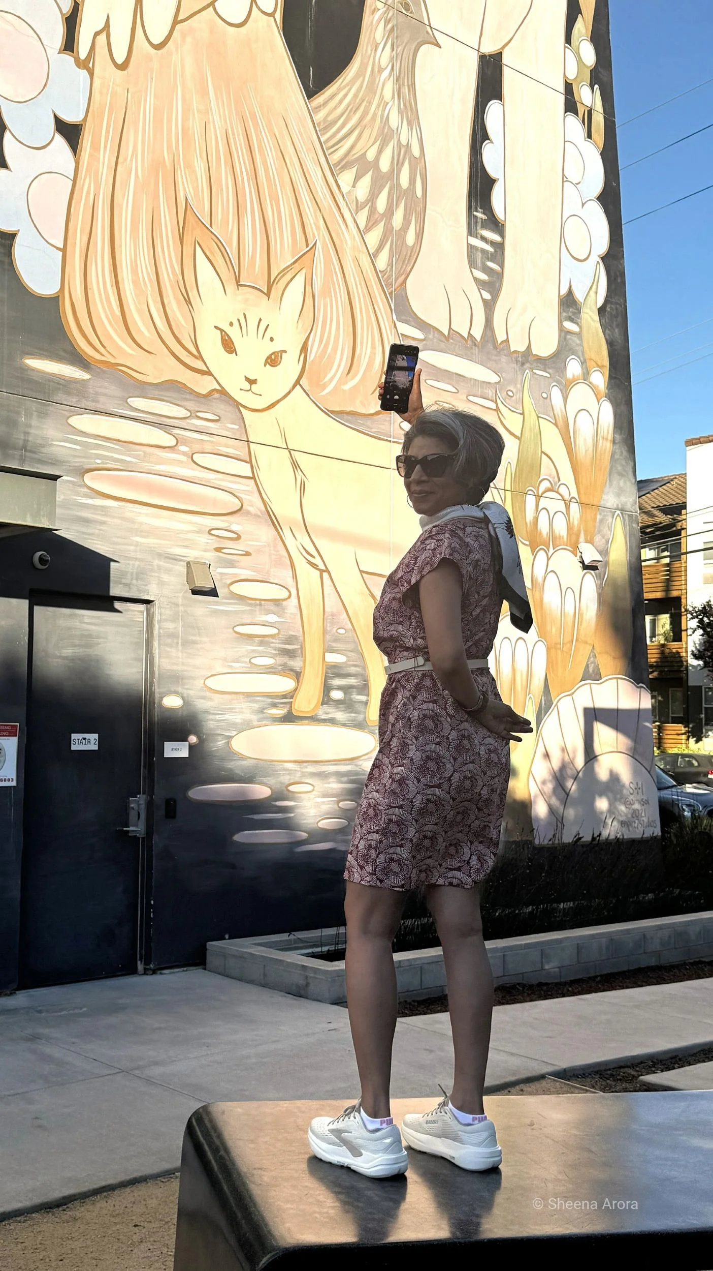 Sheena Arora standing on an outdoor table in Downtown San José to capture high-angle street art on a smartphone before it disappears.