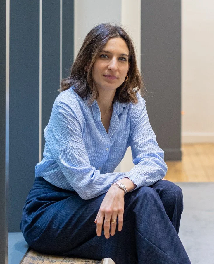 Une femme assise, portant une chemise bleue à rayures et un pantalon foncé, dans un environnement intérieur moderne.
