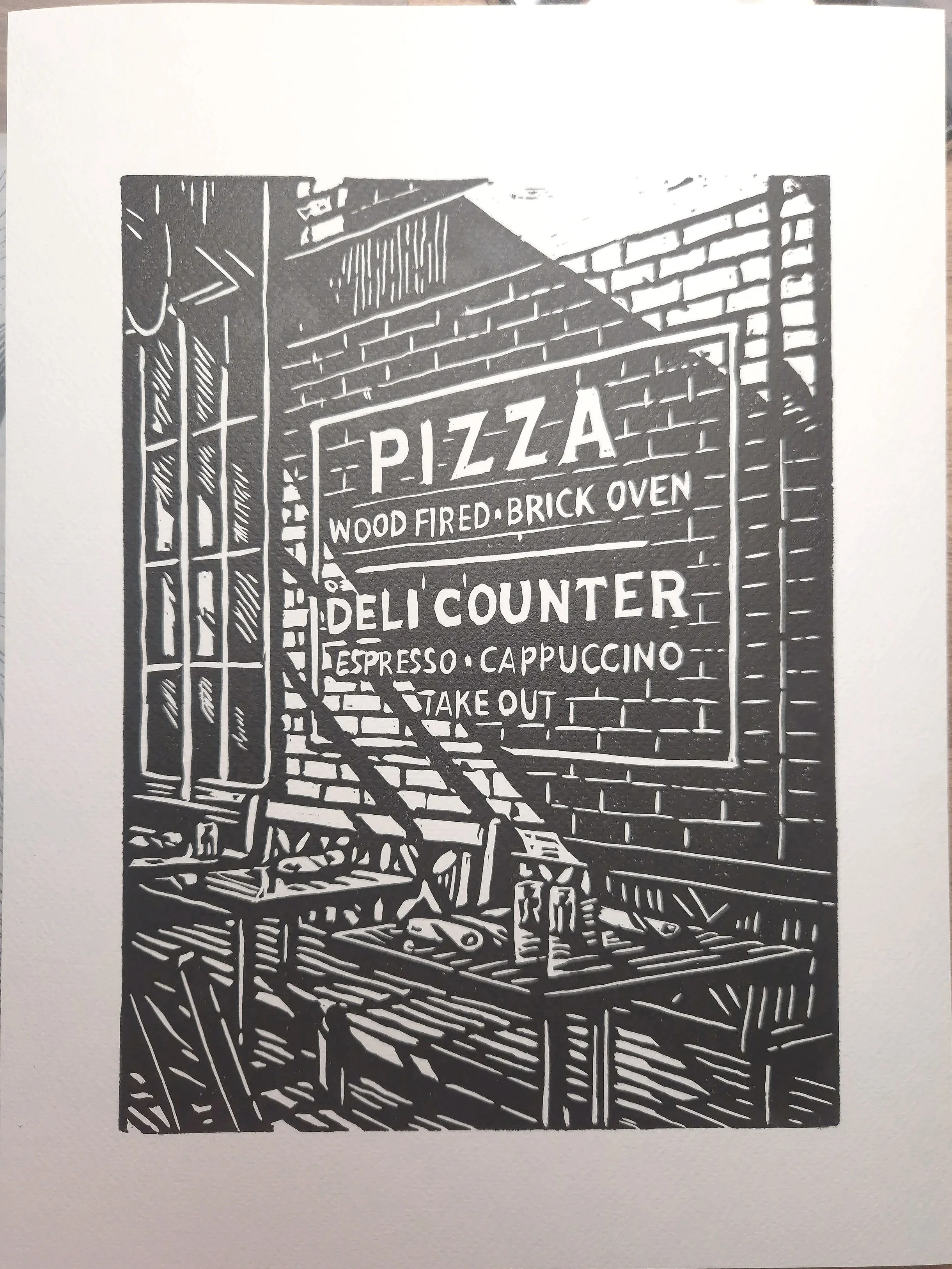 Linogravure « Caffè all’Angolo » – estampe d’art noir et blanc représentant une façade de pizzeria / deli de style new-yorkais, gravée et imprimée à la main, idéale en décoration murale.