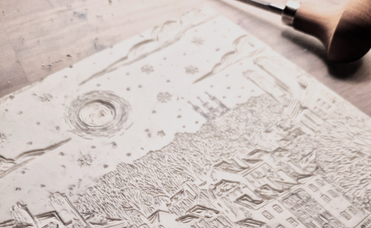 Un papier avec une gravure en relief représentant un paysage enneigé avec des maisons, des arbres et le ciel couvert de flocons de neige.