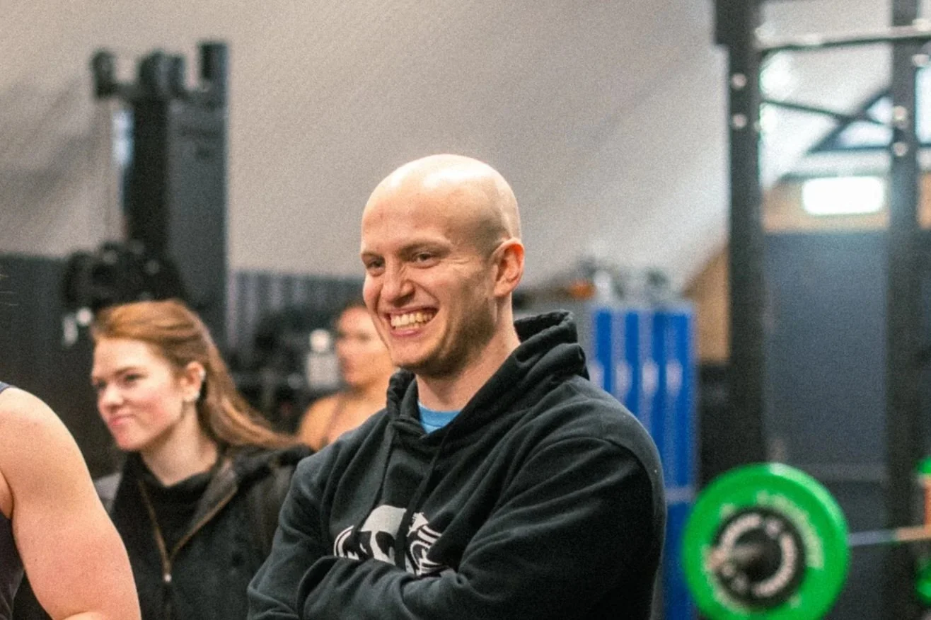 A smiling, bald man in a black hoodie at a gym, surrounded by women and gym equipment.