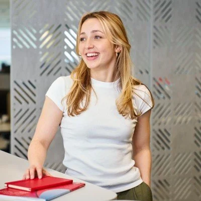 Young woman with blonde hair smiling, wearing a white t-shirt, standing by a table with red books in a modern room.