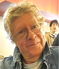 A man with curly blonde hair and glasses, wearing a denim jacket, in a room with warm lighting. There is a young man in the background.