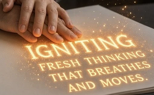 Two hand pressing firmly on the page of an open notebook, as though giving it CPR. The glowing orange text reads IGNiTING FRESH THINKING THAT BREATHES AND MOVES, surrounded by warm, sparkling light particles.