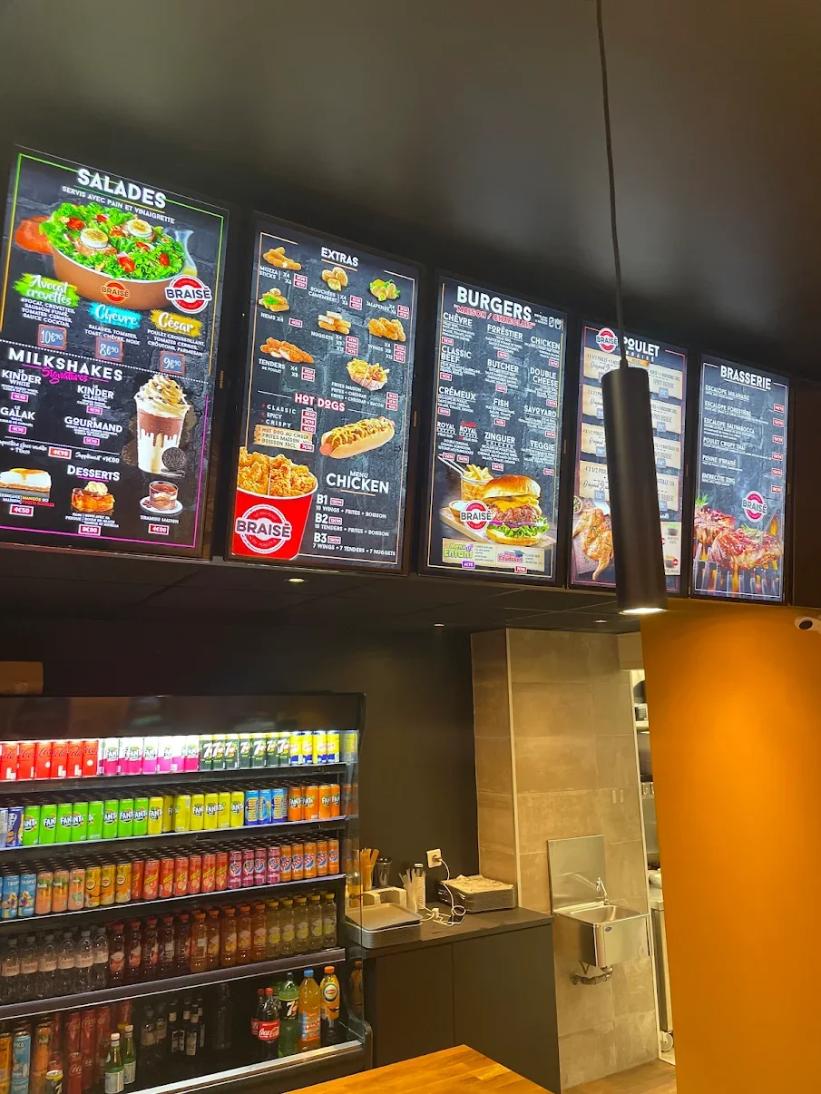 Menu numérique affichant des salades, extras, burgers, poulet frit, boissons, et desserts dans un restaurant. Réfrigérateur avec diverses boissons gazeuses et jus, comptoir avec appareils et ustensiles.