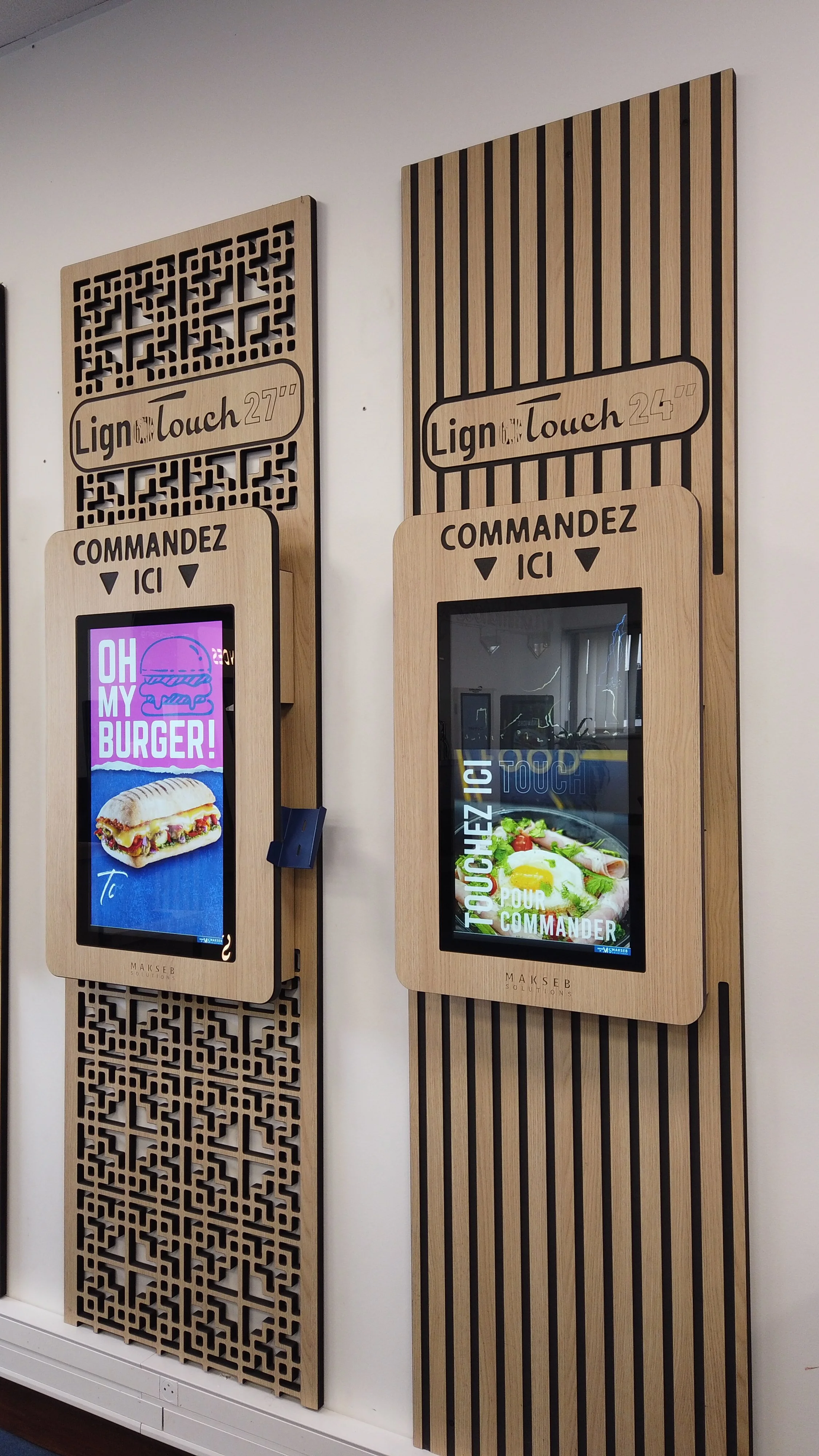 Deux bornes de commande automatisées en bois clair avec écrans tactiles, encadrées de panneaux en bois naturel, pour commander des repas, avec des inscriptions en français et des images de nourriture.
