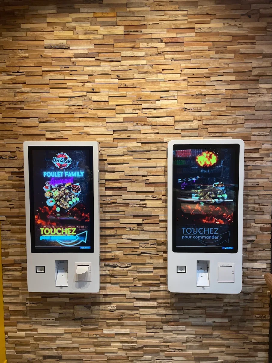 Deux écrans tactiles dans un mur en bois, avec des textes en français pour commander des aliments, probablement dans un restaurant. L'un affiche 'POULET FAMILY' et l'autre 'pour commander'.