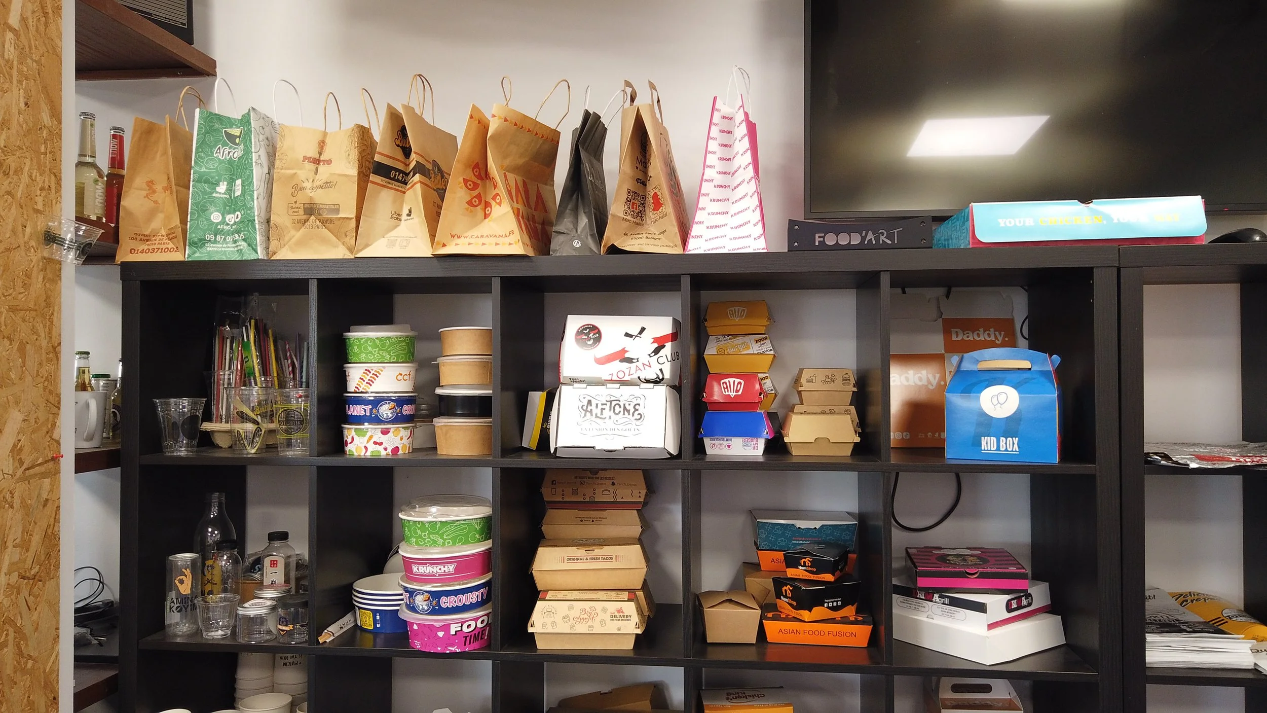 Étagère de stockage avec sacs en papier en haut, contenants en carton, ramequins, et boîtes à lunch en bas. Dispose de divers objets liés à la restauration ou à la cuisine.