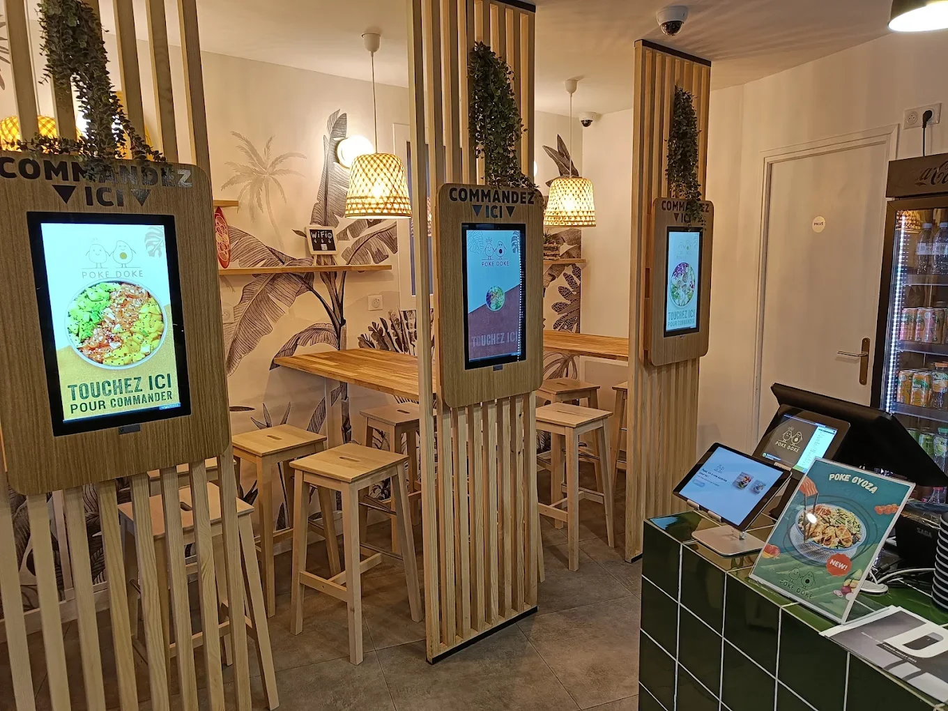 Espace de commande en self-service dans un restaurant, avec des écrans tactiles en bois, des tabourets en bois, une décoration tropicale au mur et un distributeur de boissons à droite.