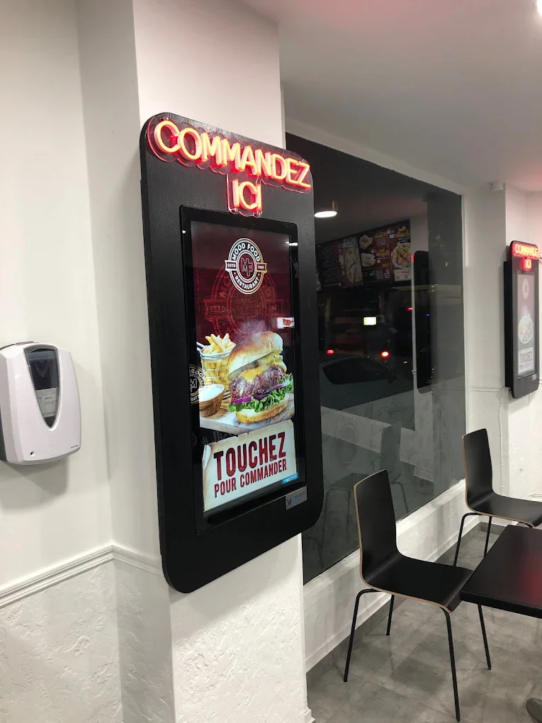 Panneau de commande pour commander de la nourriture à emporter, affichant une image d'un burger, des frites et une boisson, avec un message en français 'TOUCHEZ POUR COMMANDER' dans un restaurant de restauration rapide.