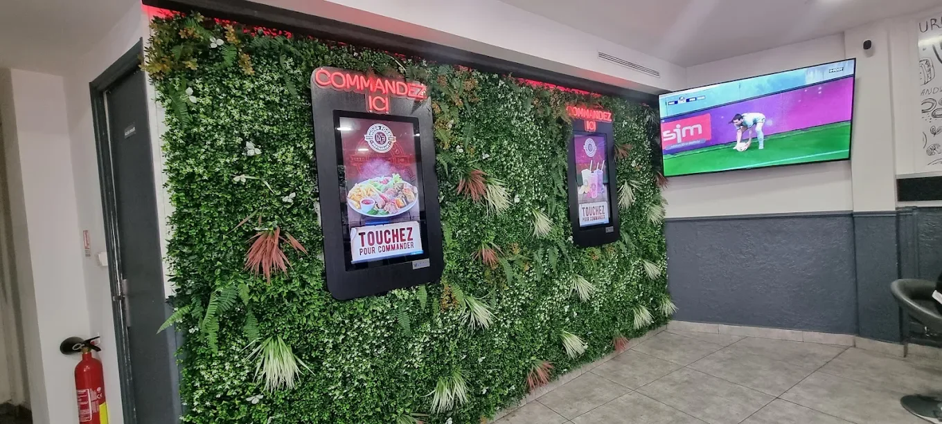 Mur végétal avec deux écrans tactiles pour commander de la nourriture et une télévision à côté montrant un enfant jouant au baseball dans un espace intérieur.