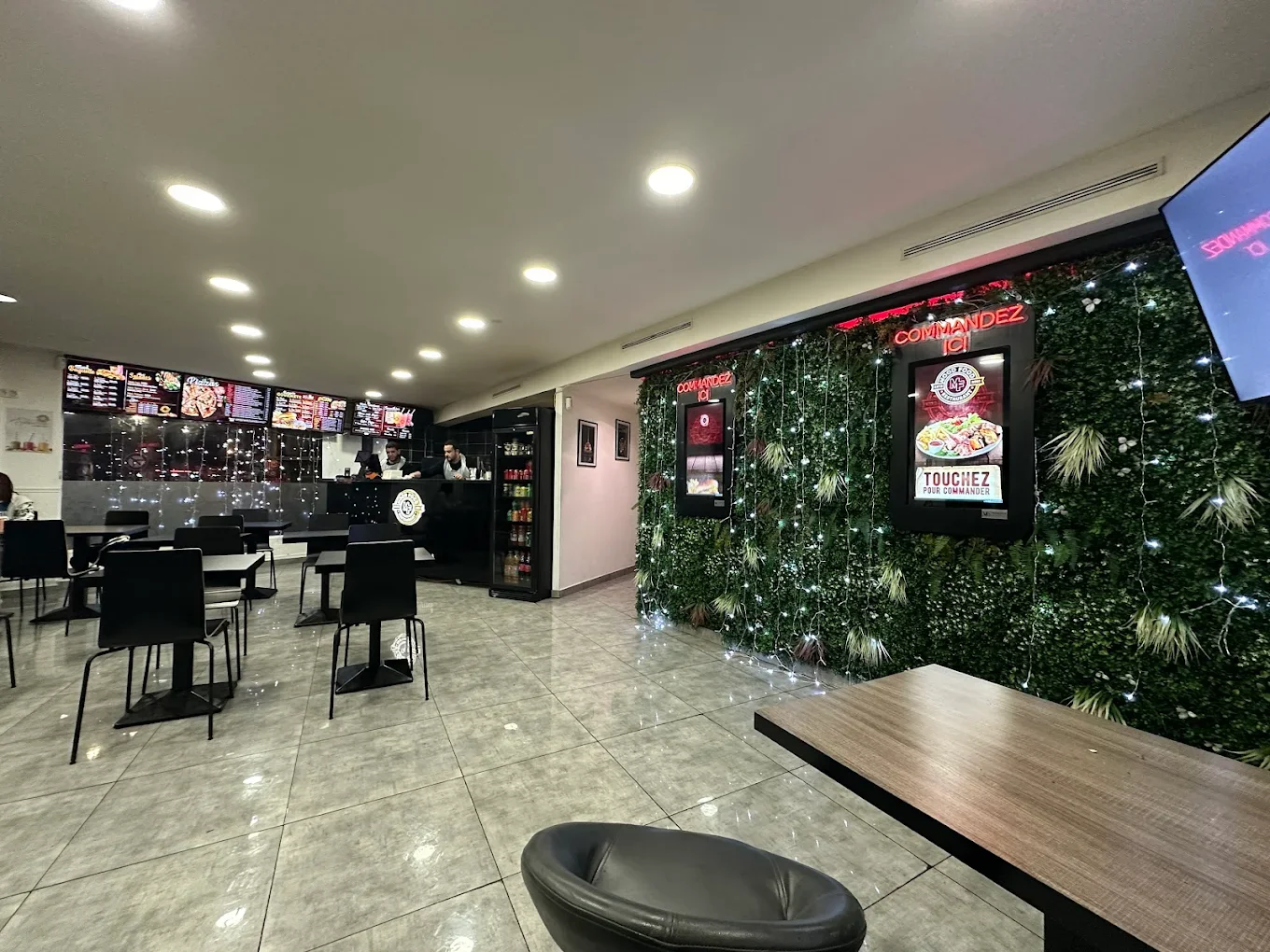 Intérieur d'une pizzeria moderne avec un espace de commande numérique, mur végétal décoratif avec affichages numériques, tables et chaises vides, comptoir avec employés et menu digital en haut.