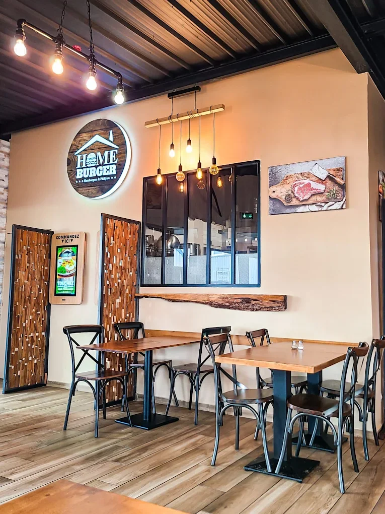 Intérieur d'un restaurant avec deux tables en bois, chaises en métal, éclairage suspendu et décorations murales, y compris un panneau lumineux 'Home Burger' et une fresque avec une pièce de viande.