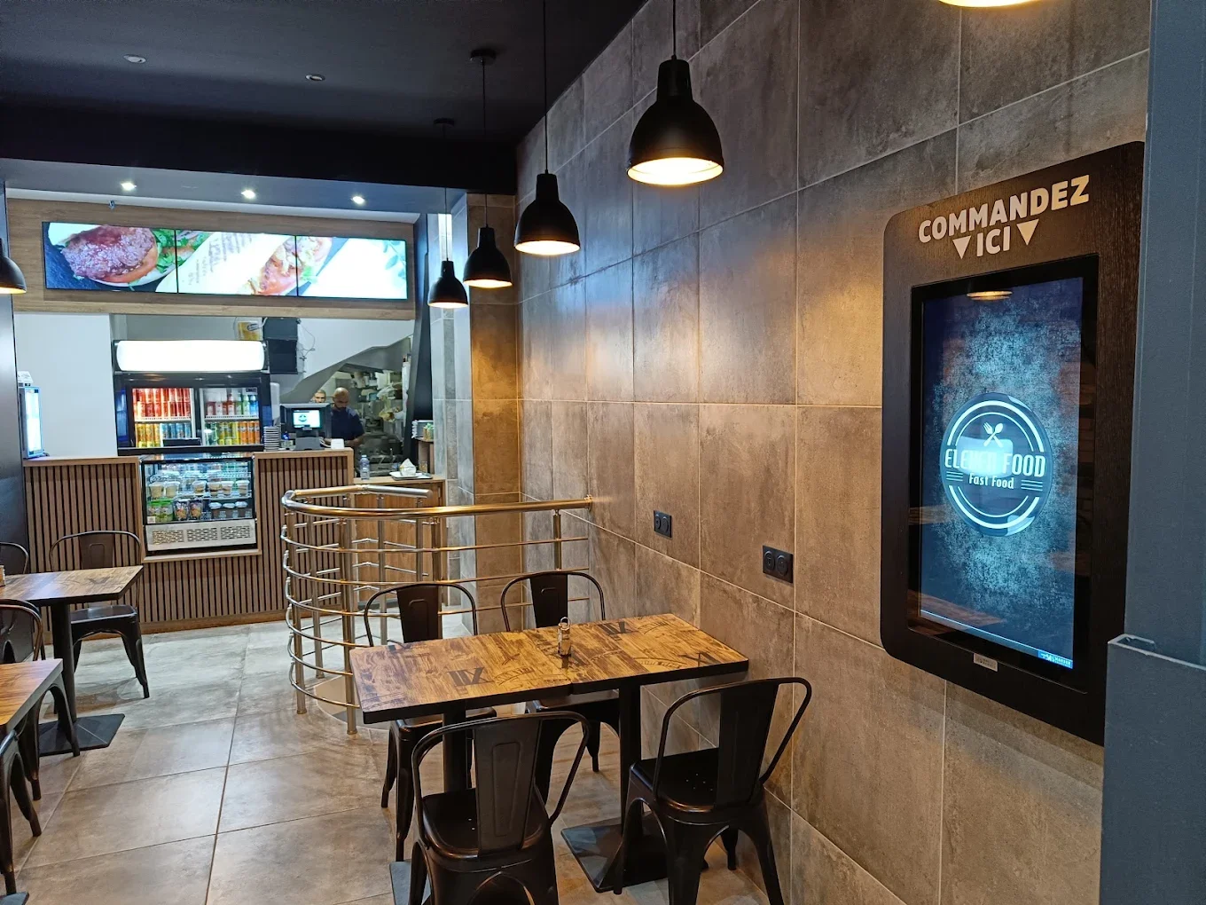 Intérieur d'un restaurant de fast-food avec tables en bois, chaises en métal, mur en carrelage, lumière suspendue noire, et un écran tactile pour commander, avec une cuisine en arrière-plan. 