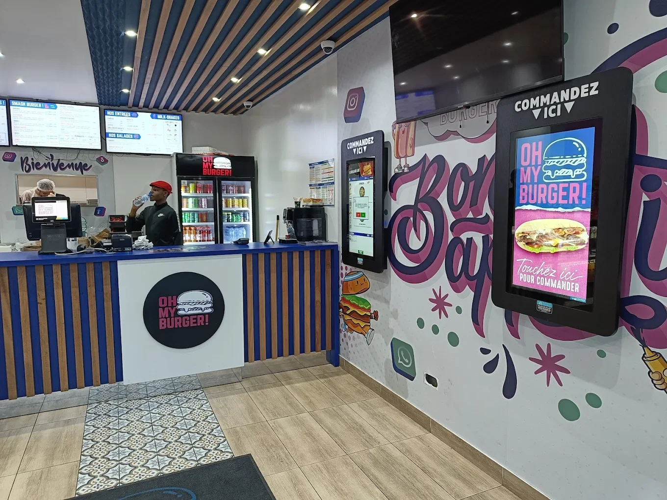 Intérieur d'un restaurant de burger avec un comptoir, un employé en uniforme, un réfrigérateur à boissons, et un mur décoré avec des écrans de commande numériques, affichant des images de hamburgers.