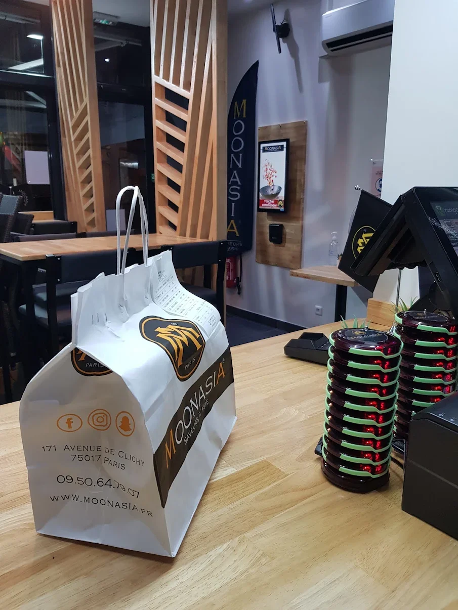 Une table dans un restaurant ou café avec un sac en papier blanc portant le logo de MoonAsia, un magasin ou restaurant asiatique, ainsi que plusieurs présentoirs de cartons noirs empilés en forme de pyramide. En arrière-plan, il y a une bannière avec