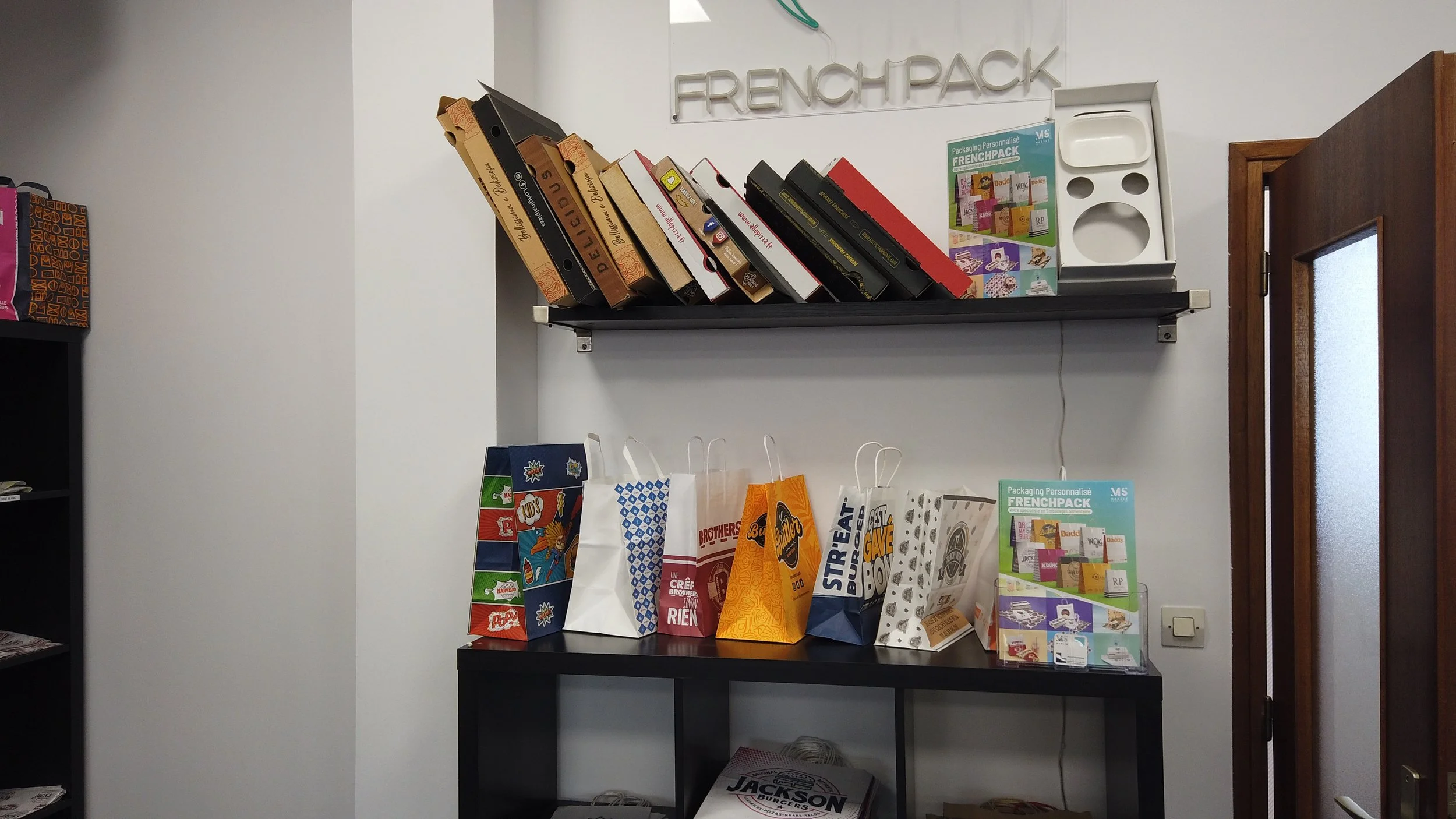 Espace de bureau ou de boutique avec des étagères contenant des dossiers, des sacs en papier de différentes couleurs et un pamphlet promotionnel. La porte en bois est visible à droite.