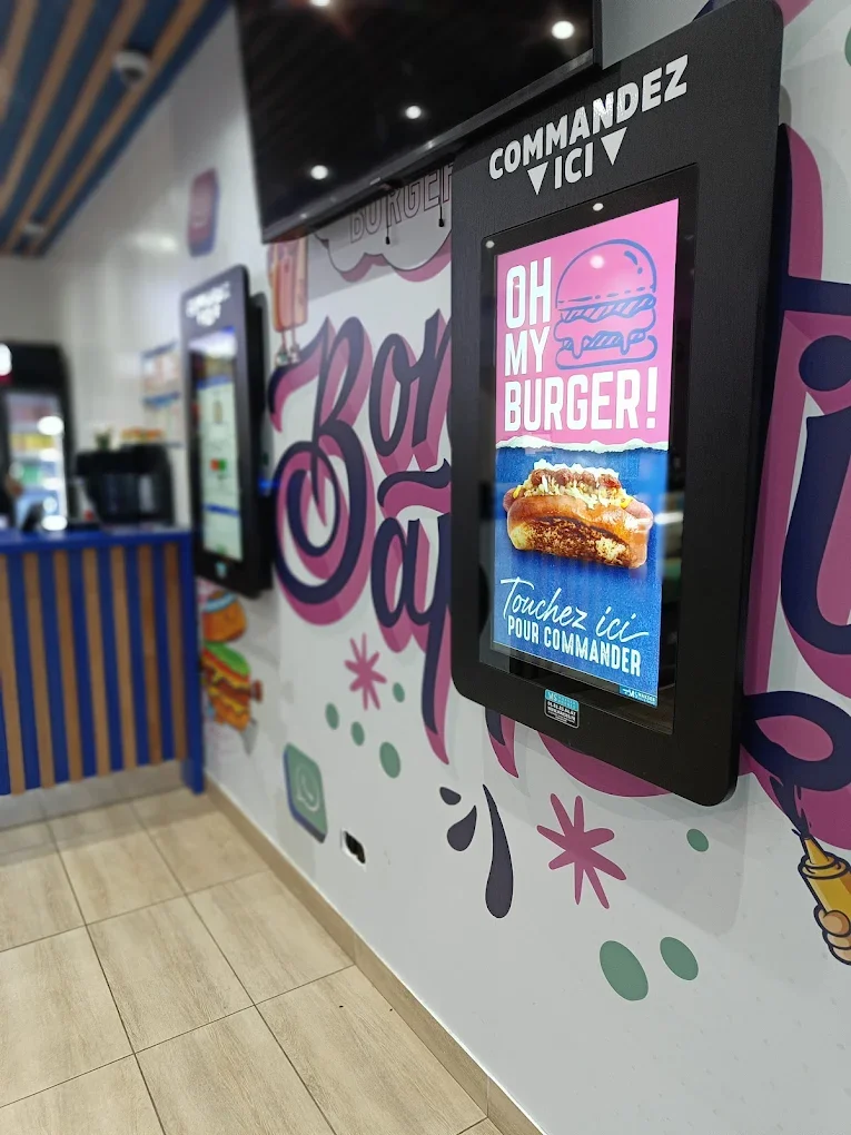 Un mur avec une grande enseigne colorée et un écran interactif pour commander un burger dans un restaurant rapide. L'enseigne sur l'écran affiche 'OH MY BURGER!' et une image d'un burger.