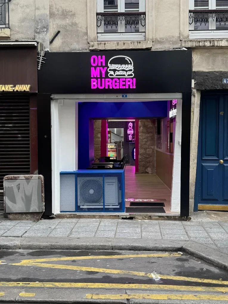 Façade d'un fast-food avec enseigne lumineuse 'OH MY BURGER!' en violet, entrée menant à un intérieur avec des sièges et un éclairage rose pink, décor néon avec des hamburgers et une glace.
