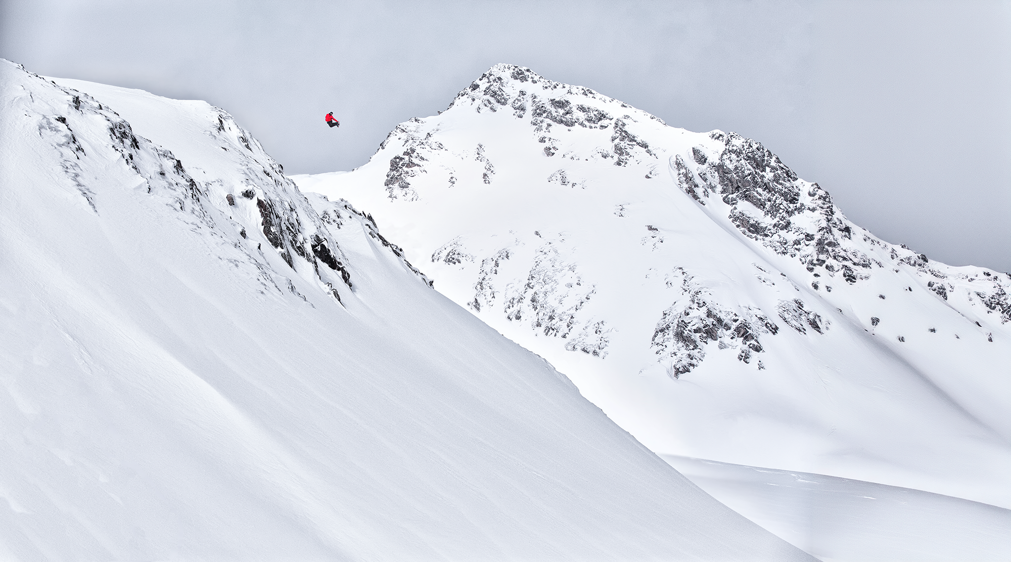 Ein Skifahrer springt in die Luft vor einer schneebedeckten Berglandschaft mit bewölktem Himmel.