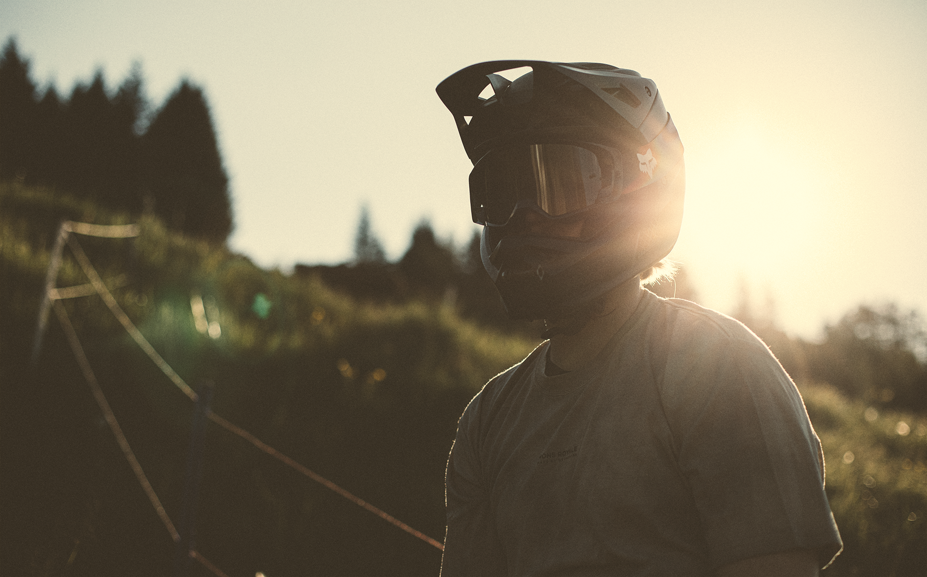 Person mit Motorradhelm im Sonnenlicht, im Freien auf einem Hang mit Seilspannern im Hintergrund.